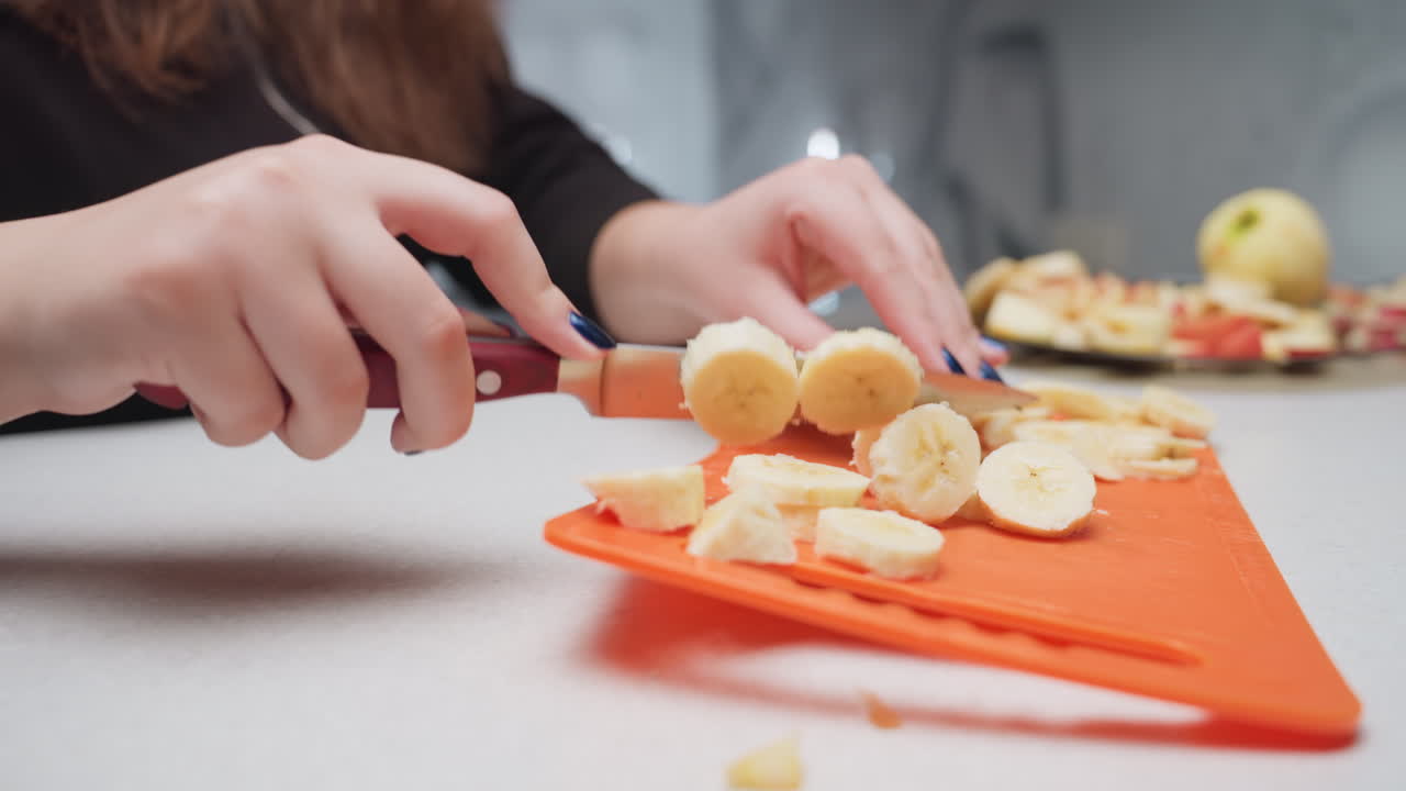 Close up manicured hands dicing plantain with knife on orange rubber board, round slices gathering on surface, plate with apple pieces visible in background, kitchen prep scene, fresh fruit snack