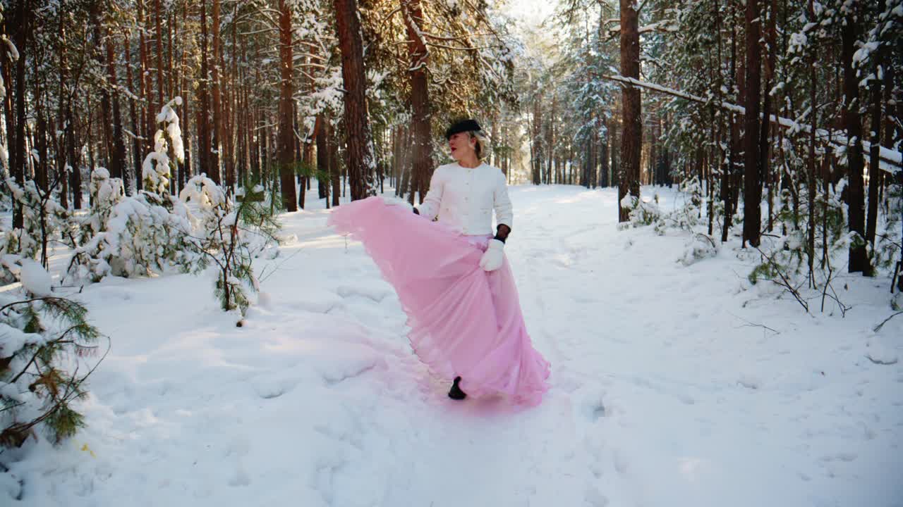 Elegantti nainen vaaleanpunaisessa mekossa kävelemässä ja pyörähtelemässä lumisessa metsässä