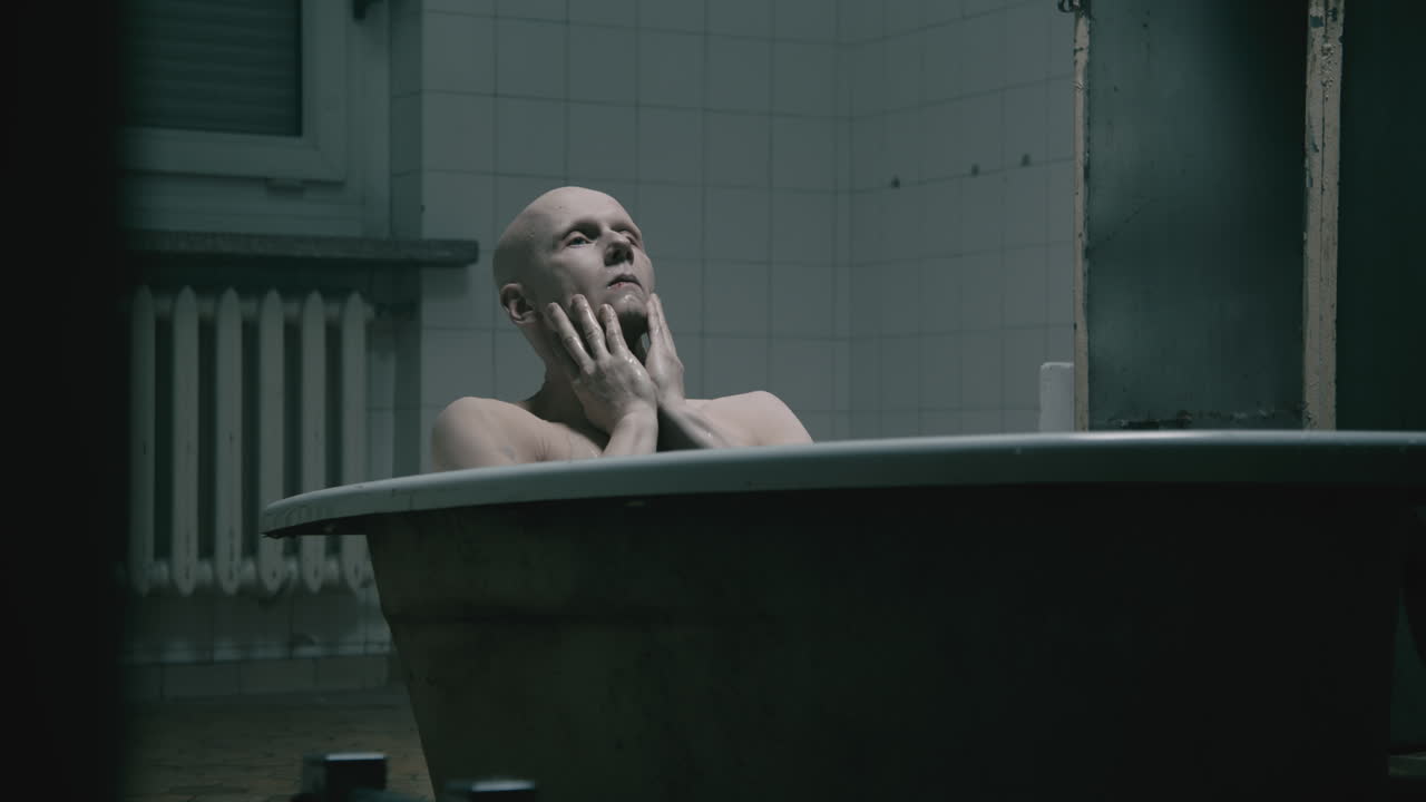 Pale bald skinny man takes a bath in the creepy old bathroom at night