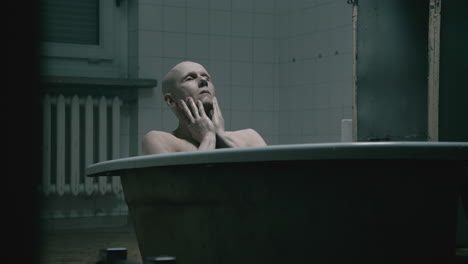 pale bald skinny man takes a bath in the creepy old bathroom at night