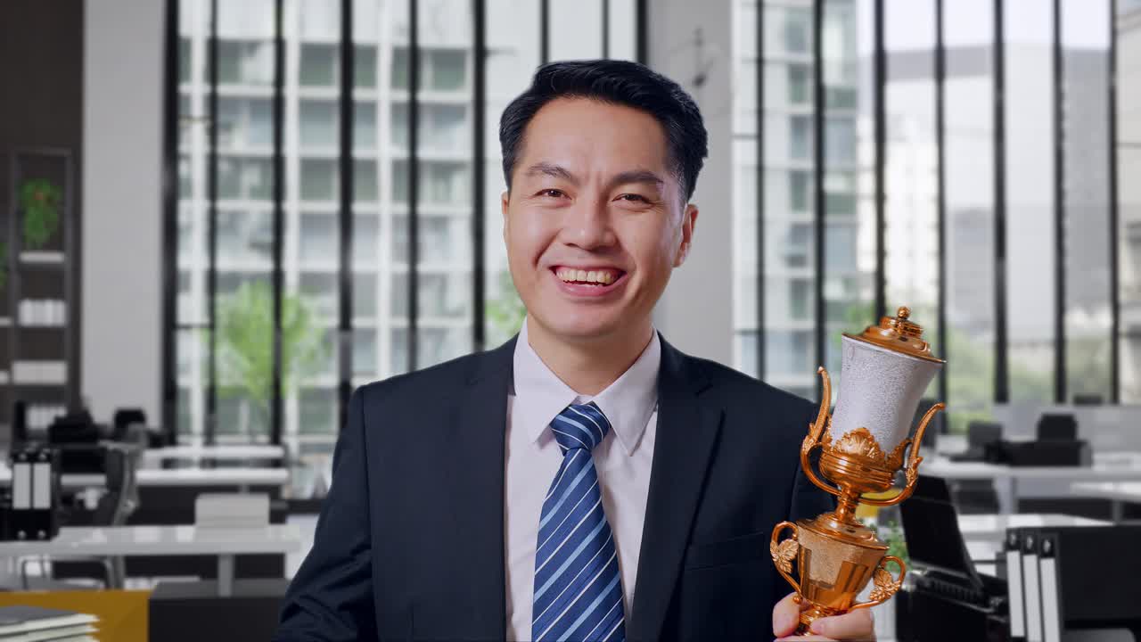 Close Up Of Asian Business Man In A Suit And Tie With A Gold Medal And Trophy Showing Peace Gesture And Smiling To Camera As The First Winner In Office