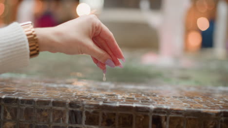 poised hand in white sweater gently touching water surface of mosaic tiled mall fountain under bokeh lighting, subtle ripples spreading outward, blurred jets in background evoke calm serene mood