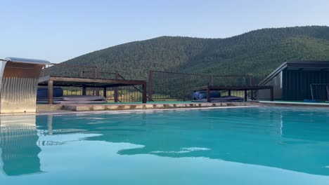 outdoor swimming pool with crystal clear water surrounded by a fence with mountains and pine forest in the background, sunset