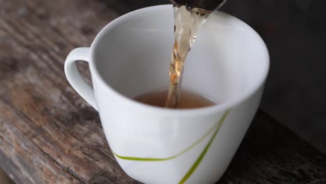 Toma-En-Cámara-Lenta-De-Verter-Una-Taza-De-Té-En-Una-Taza-Blanca-Sobre-Una-Mesa-De-Madera