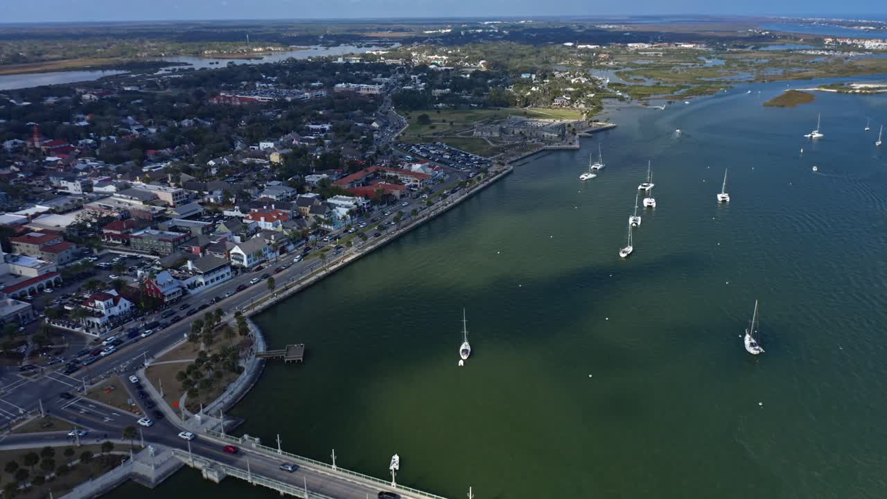 Area urbana costiera con un ponte che attraversa l'acqua e barche a vela ancorate vicino ai moli, circondata da strade cittadine, edifici e spazi verdi lungo la costa in un ambiente temperato