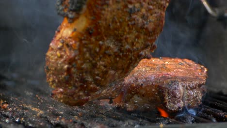 a pair of meat tongs turn a nearly cooked juicy rib eye steak on a grill and presses on it as flames shoot up