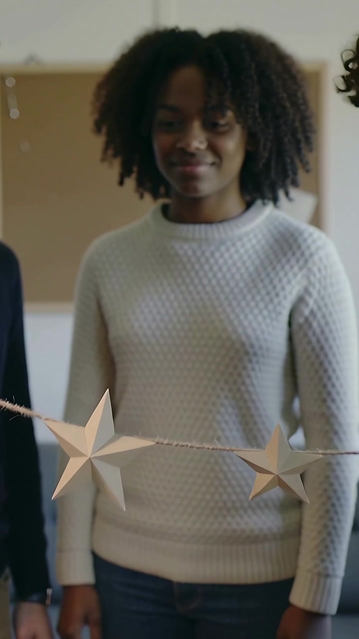 Vertical video: Reaching hands adjusting paper star garland at home, woman in cream knit decorating