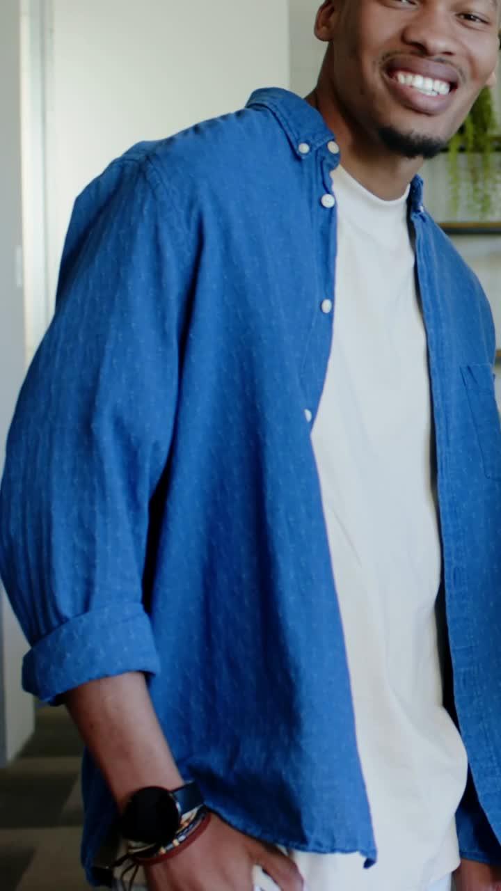 Video verticale: Un uomo in piedi con camicia blu e orologio, che si gira sorridendo vicino alla porta dopo aver notato qualcuno.
