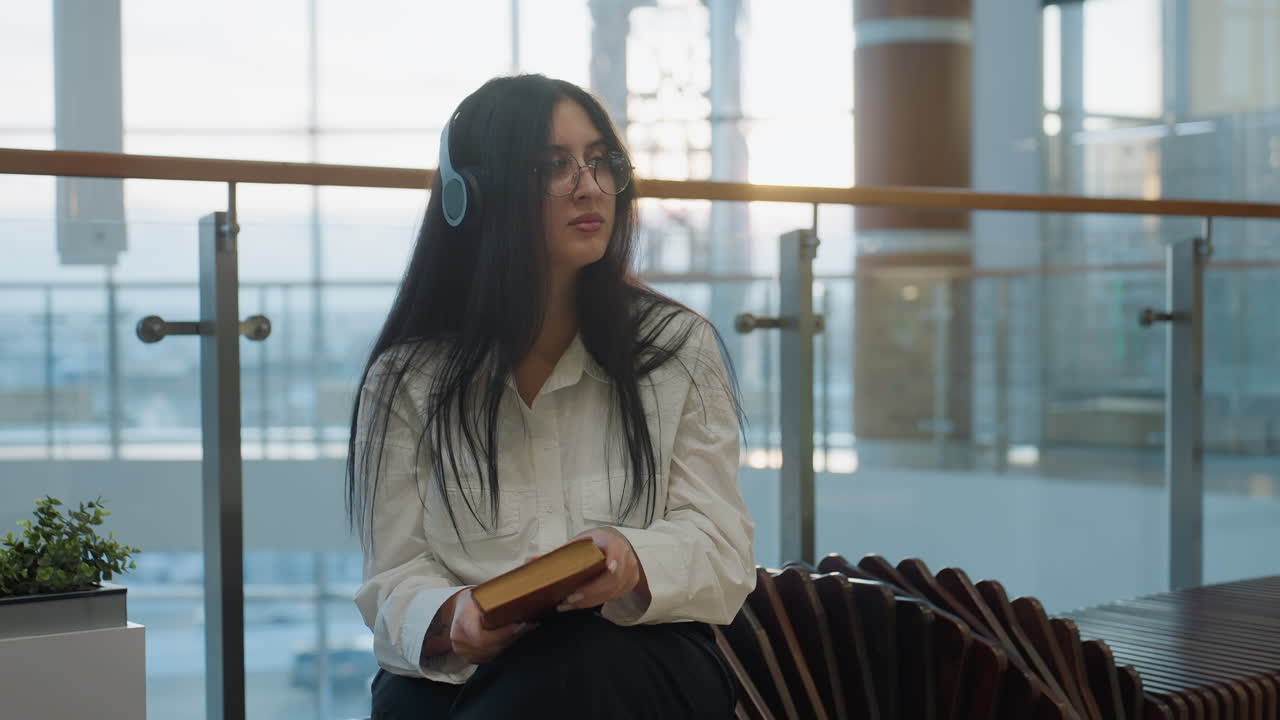 Woman with long dark hair closes book and gazes upward thoughtfully while seated indoors wearing glasses and headphones, surrounded by soft light and modern interior with glass railings