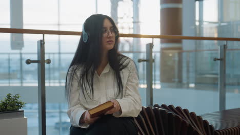woman with long dark hair closes book and gazes upward thoughtfully while seated indoors wearing glasses and headphones, surrounded by soft light and modern interior with glass railings