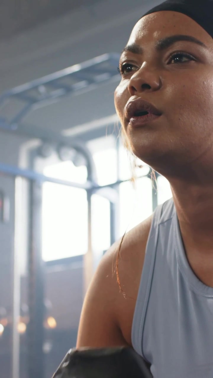 Vertical video: Lifting woman inhaling, raising black dumbbell at gym wearing blue top for strength