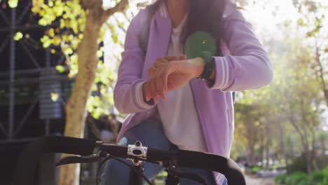 Asiatische-Frau-Benutzt-Smartwatch,-Während-Sie-Im-Park-Auf-Dem-Fahrrad-Sitzt