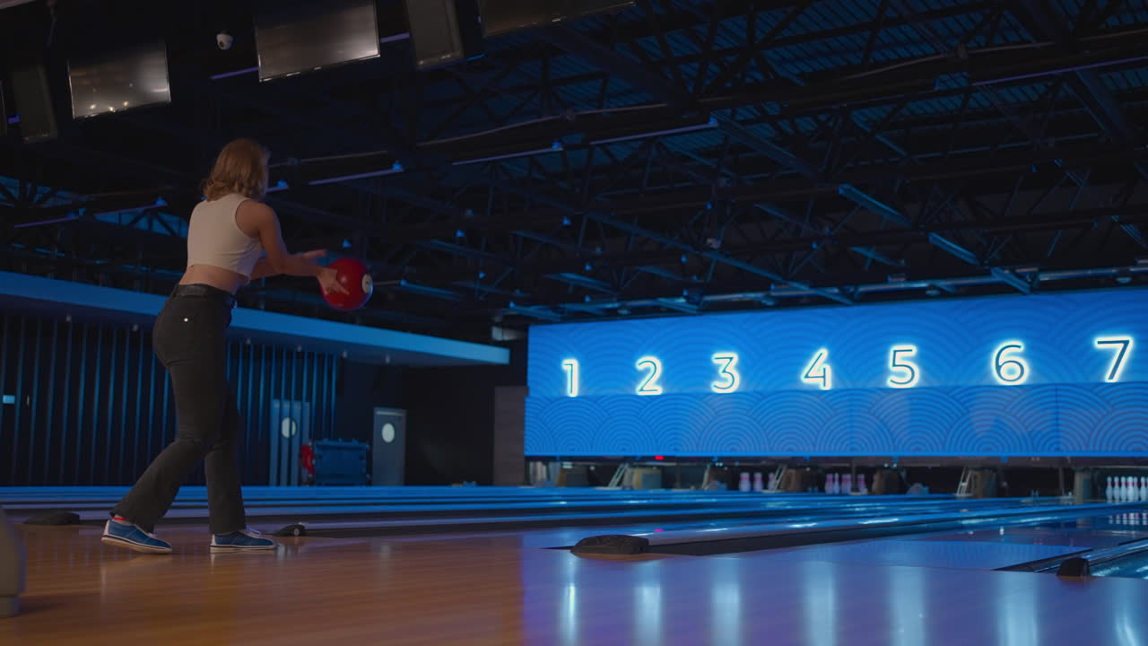 A blonde woman in bowling throws a ball in slow motion and knocks down skittles. Play bowling. Throw balls on the floor of the bowling club