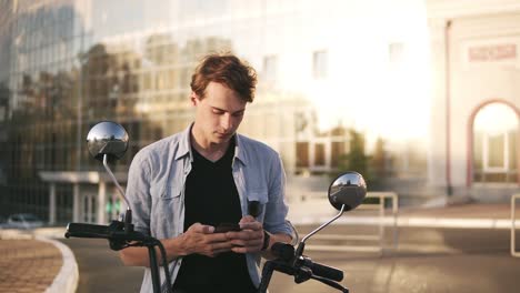 retrato al aire libre de un joven moderno con teléfono móvil en la calle, sentado en una minibike. paredes de espejo modernas construyendo en el fondo