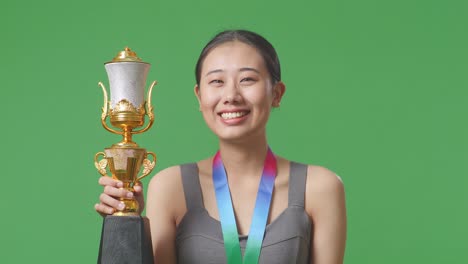 close up of asian woman wears a gold medal and holds a gold trophy in her hand as the first winner smiling to camera on green screen background in the studio