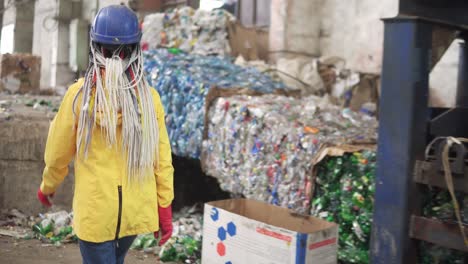 Woman-worker-with-dradlocks-in-yellow-and-transparent-protecting-glasses,-hard-hat-and-mask-working-with-equipment-on-recycle-plant.-Press-machine-with-used-plastic-bottles-and-differen-plastic-garbage.-Footage-of-automized-process