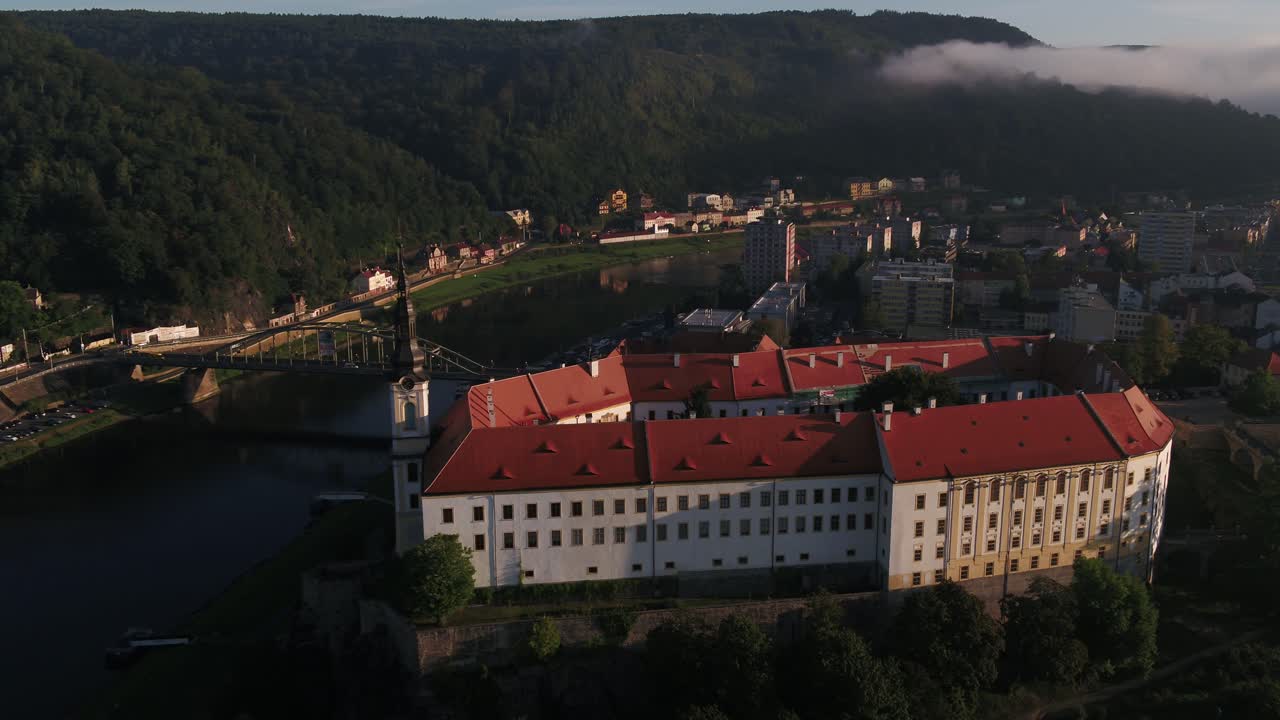 Uma ampla tomada de drone revela o Castelo de Decin e o Rio Elba cercados por névoa e luzes da cidade durante a hora dourada.