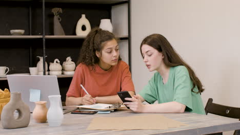 pottery studio owner talking with a client and taking notes on a clipboard while client showing products on mobile phone 1