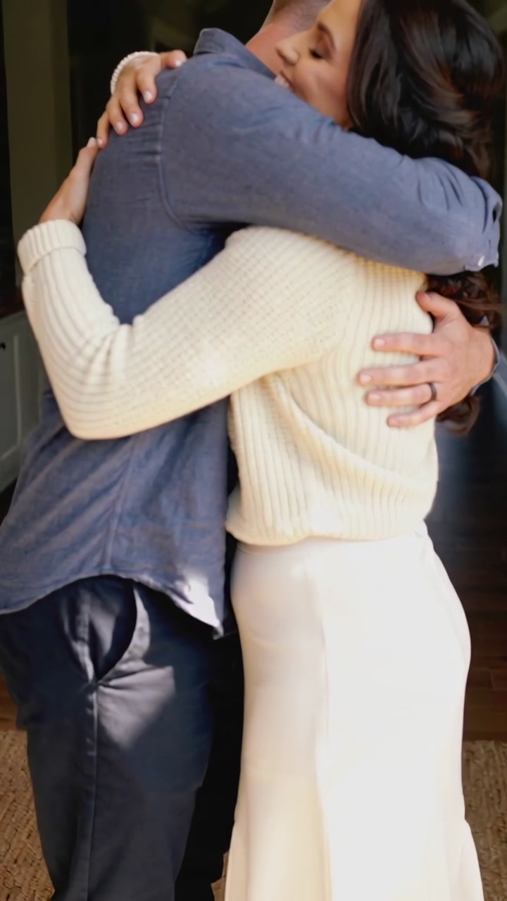 Vertical video: Hugging couple embracing and pressing heads in hallway, showing ring, cream sweater