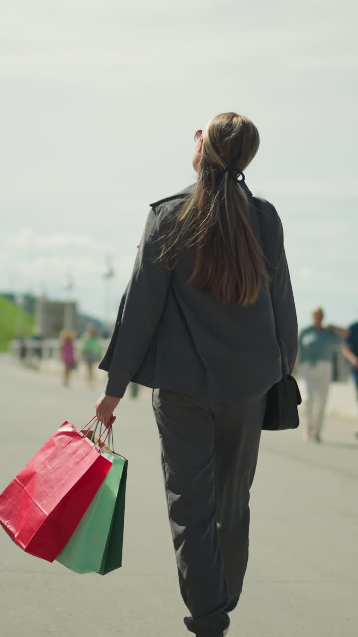 선글라스를 입은 여자가 빨간색, 민트색, 녹색 쇼핑백을 들고 언덕을 향해 부드럽게 흔들고 있으며, 배경에는 반대 방향에서 다가오는 흐릿한 사람들이 있습니다.