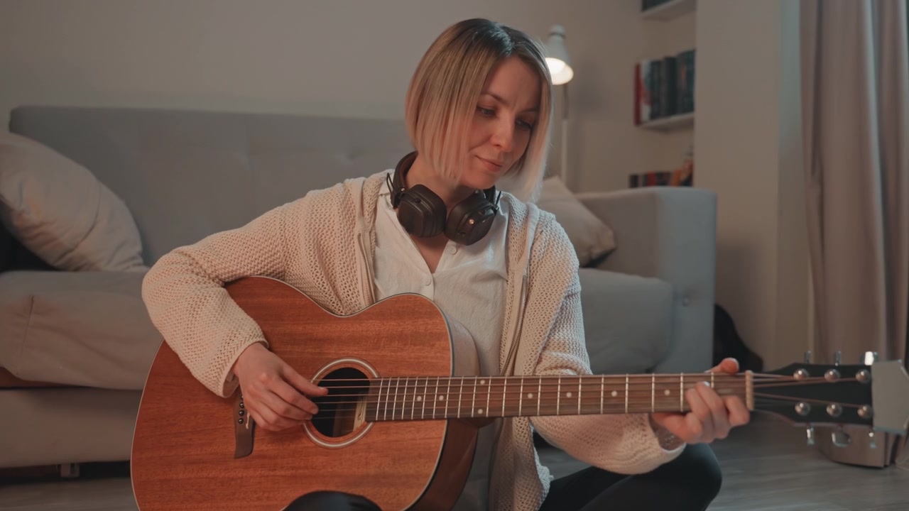Donna bianca che strimpella una chitarra acustica a terra con le cuffie, soggiorno luminoso e accogliente, sorriso giocoso e ritmo energico, sessione di prove e pratica casalinga vivace, momento creativo spontaneo.