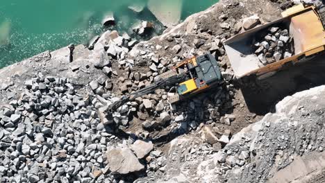 aerial top view of an excavator loading stones into a truck by a quarry lake