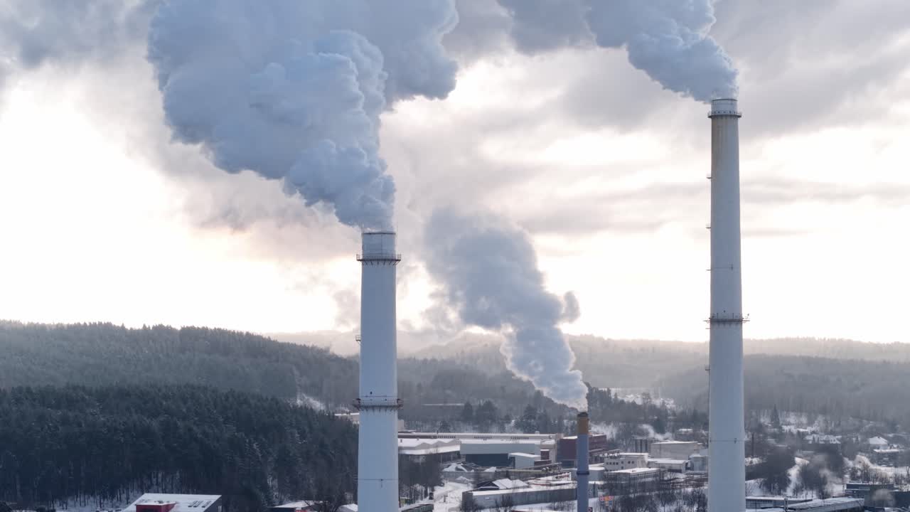 Vista aerea da drone con teleobiettivo delle ciminiere della centrale termoelettrica di Vilnius che rilasciano un denso vapore sopra la foresta innevata e il margine della città, mettendo in mostra l'infrastruttura di riscaldamento del distretto e i problemi relativi alla qualità dell'aria.