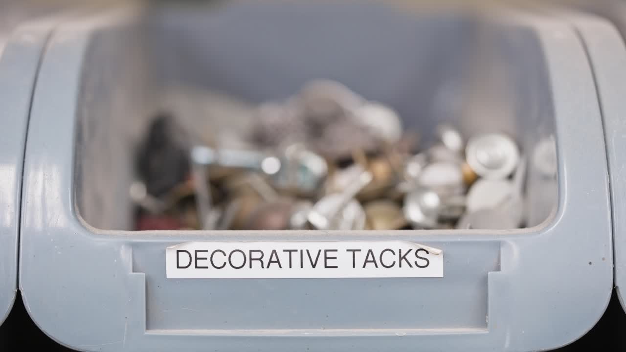 Rack focus reveals 'decorative tacks' label on bin filled with metal tacks
