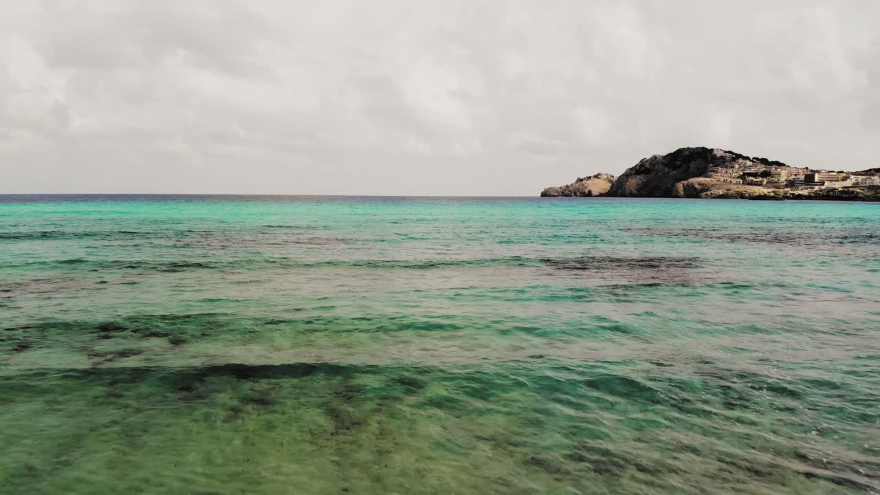Low aerial drone shot panning backwards of clear blue water of the Mediterranean at a beach in Majorca with a town in the distance nested in the hills.
