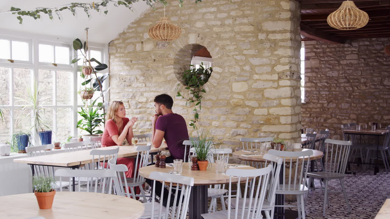 una pareja de adultos jóvenes de raza mixta sentados en una mesa tomados de la mano y hablando en un restaurante vacío