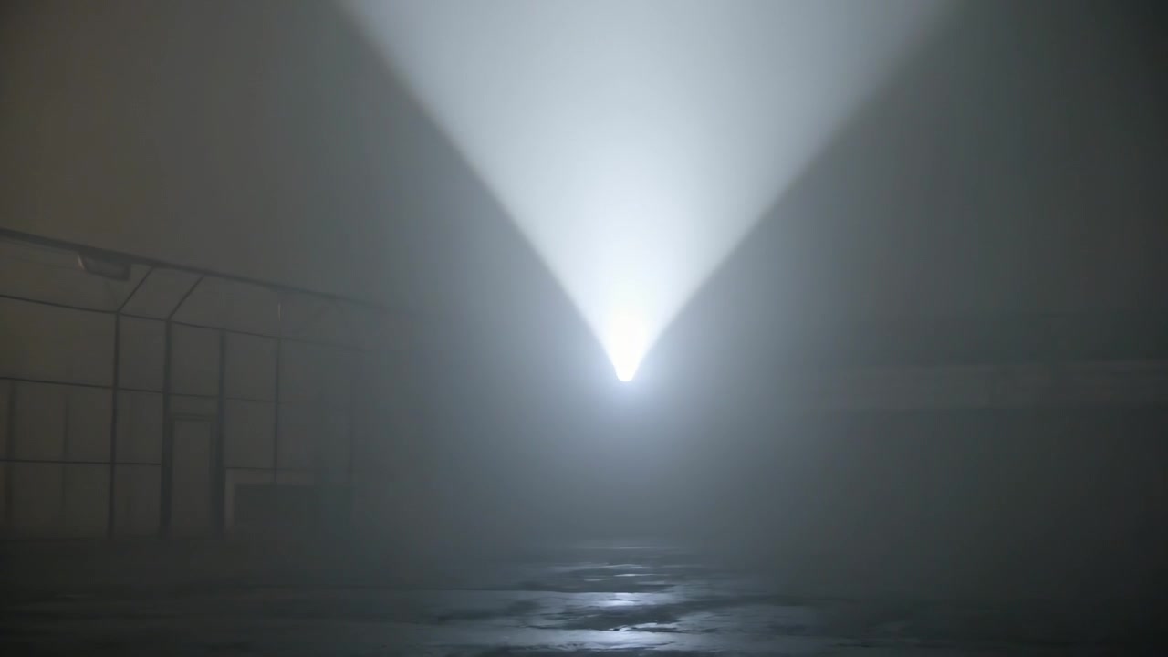 Holofote aceso criando um feixe varredor e que se alarga através de uma baía nebulosa, iluminando o chão molhado e uma parede de metal