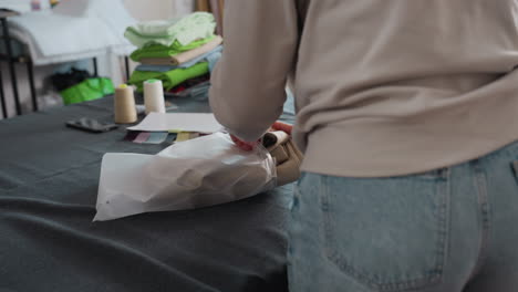 garment maker folds beige clothing piece then slides item into transparent nylon packaging bag on cutting table inside bright sewing workshop, demonstrating careful handling technique
