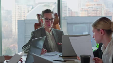a confident middle-aged blonde girl with glasses and a business gray suit speaks to her businesswoman colleagues while sitting at a table with laptops and important papers in the office