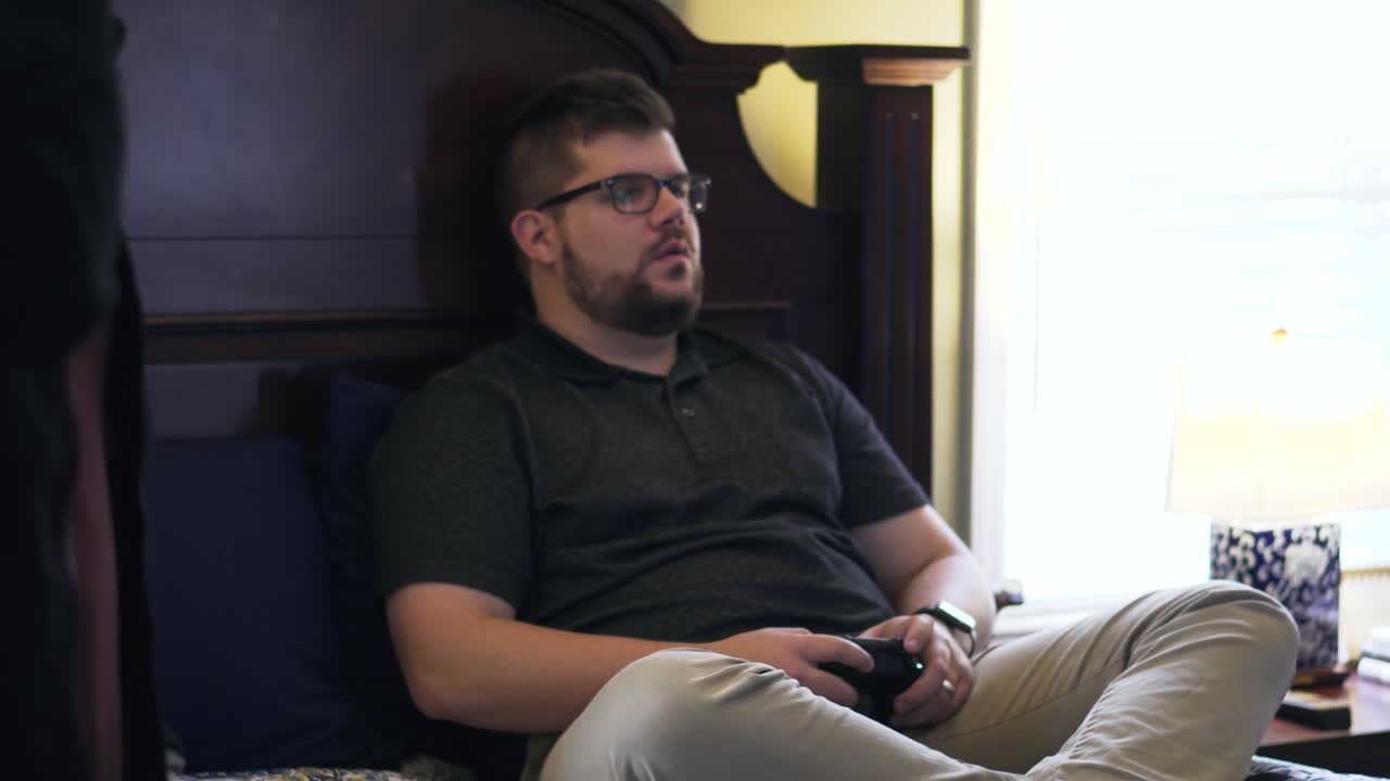 Man laughs and tries to concentrate on a video game while his friends are talking and walking around the room