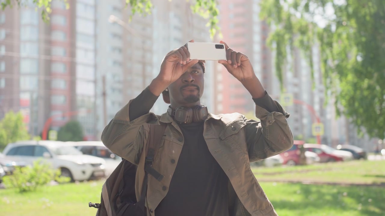 Primo piano di un uomo che tiene uno smartphone e scatta una foto in un parco urbano con alberi verdi, auto parcheggiate ed edifici alti. L'uomo indossa un berretto marrone, una giacca sopra una camicia nera e ha le cuffie al collo