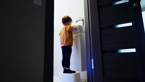 toddler boy stands on the prop near the sink in the bathroom. kid opens the faucet and watching the water flow. low angle view.