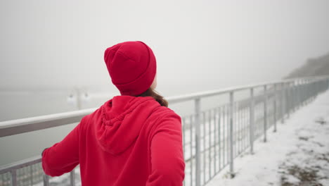 close-up of athlete exercising outdoors during winter, while stretching her leg back and forth, surrounded by snowy ground, foggy atmosphere, distant river, and light poles in background