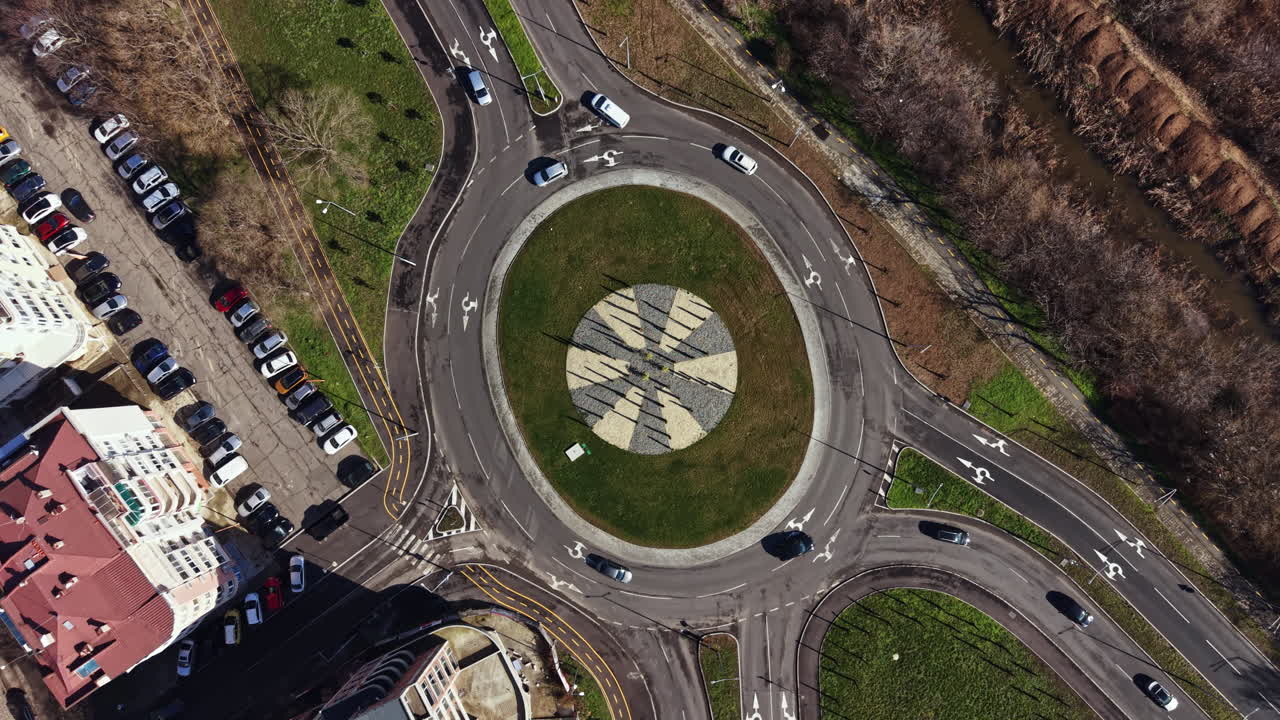 Cars move in a roundabout near a park with green grass and trees while a river runs beside the road during a sunny day