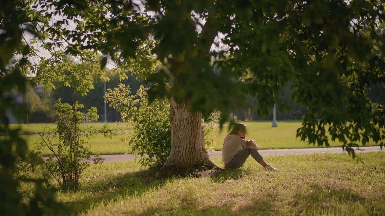 한 여인이 잔디에 앉아서 손을 다리 위에 고, 그늘진 나무 아래, 초록색으로 둘러싸여, 빛이 잎을 통해 필터링, 경로, 그리고 잔잔한 풍경이 배경입니다.