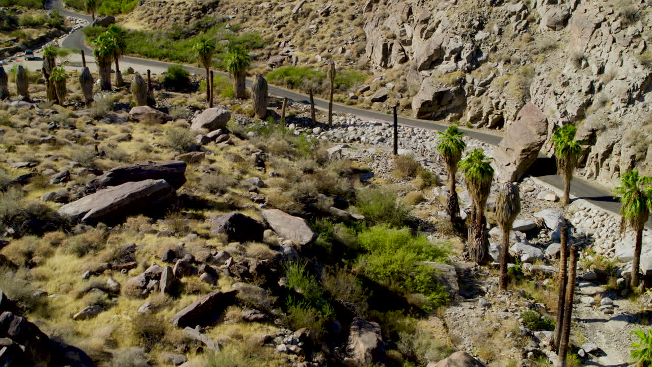 Aerial view of rugged Palm Springs mountain terrain next a road, showcasing dramatic ridges, rocky slopes, and desert textures under a clear blue sky. A striking natural landscape from above