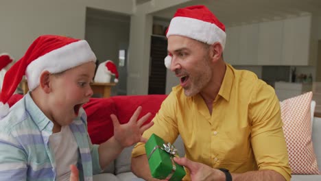 Kaukasischer-Vater-Mit-Weihnachtsmütze,-Der-Seinem-Sohn-Zu-Hause-Ein-Weihnachtsgeschenk-Schenkt