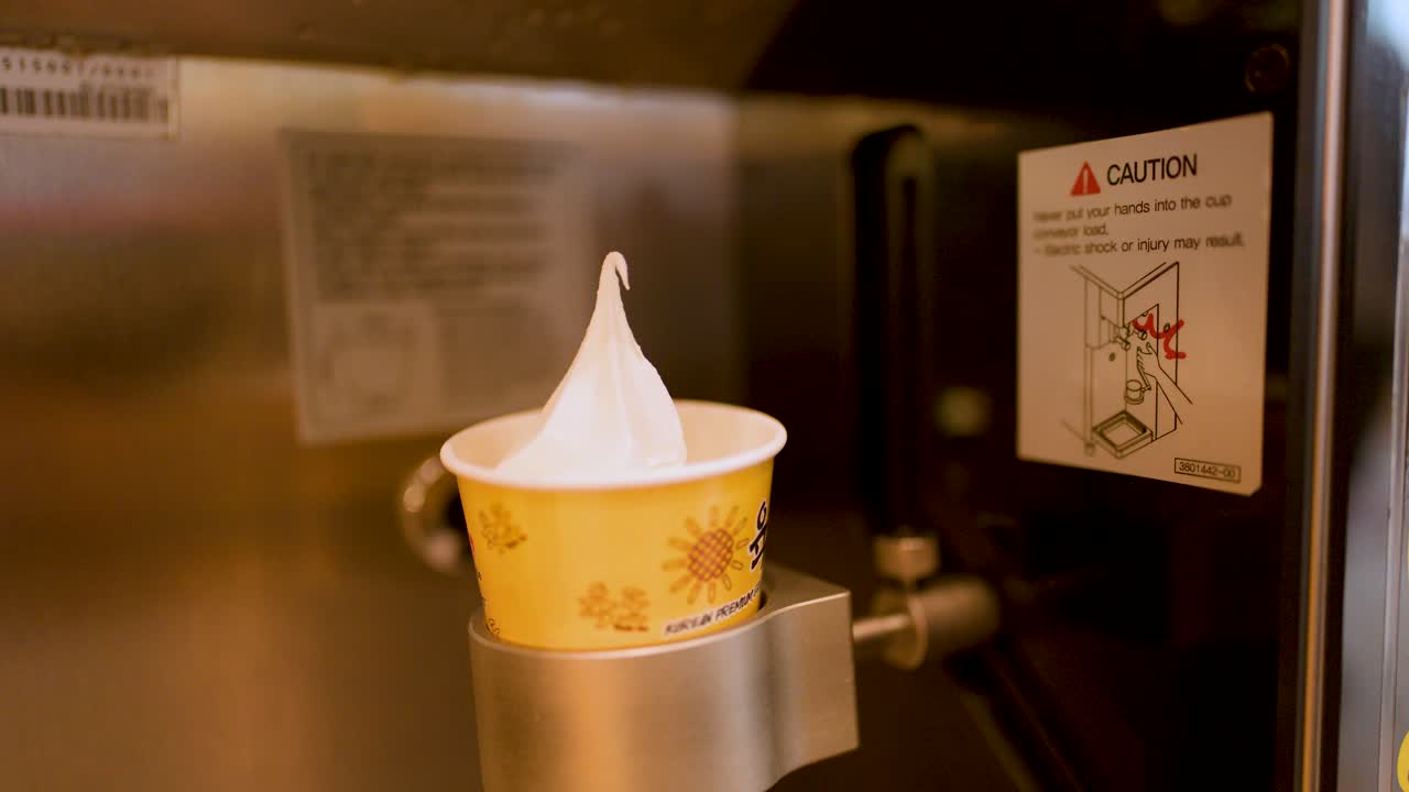 Automatic machine dispenses soft serve ice cream into cup; hand retrieves dessert under warm lighting