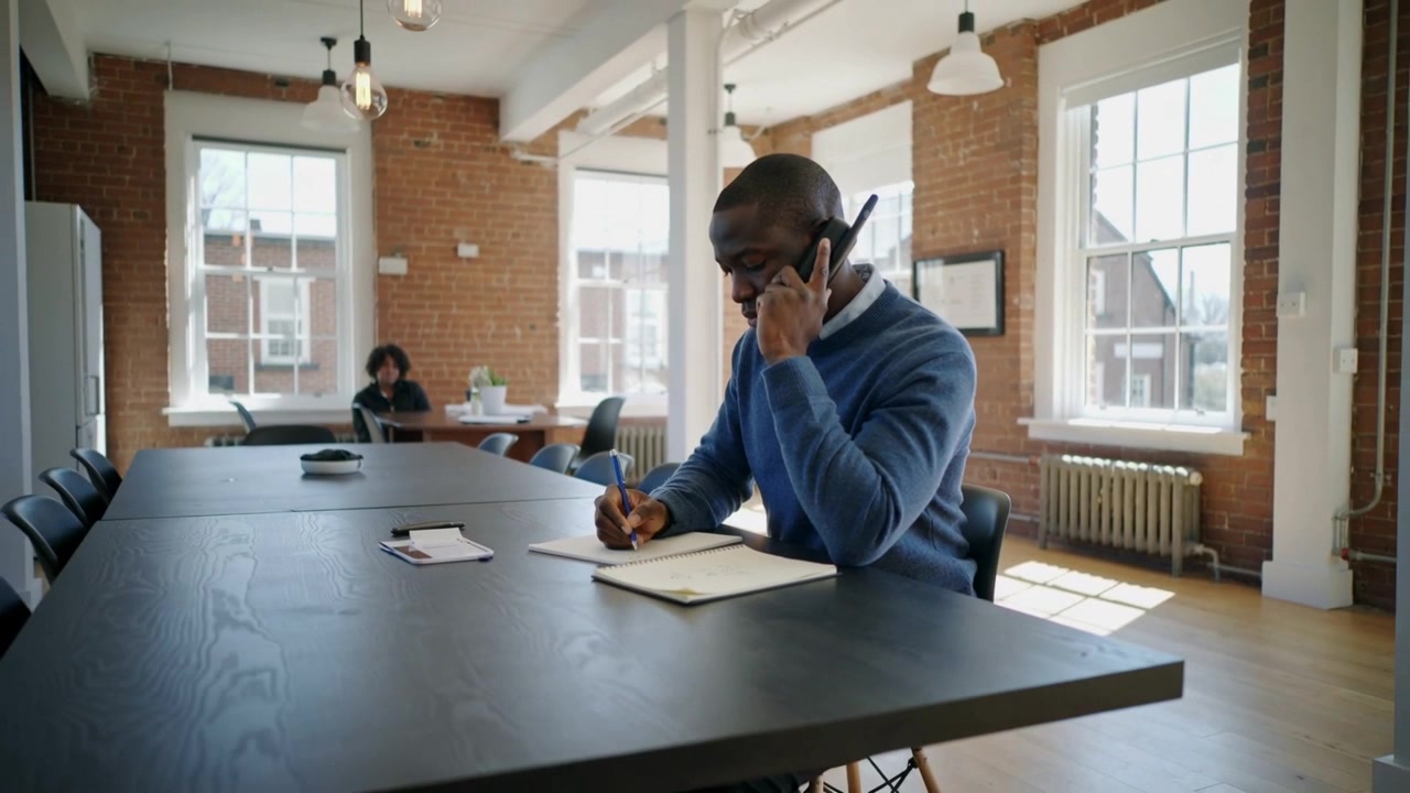 Answering call man in blue sweater holding phone taking notes in spiral-notebook at loft