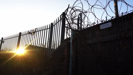 Sunset-by-razor-wired-fence,-barbed-wire-and-security-gate-at-an-old-abandoned-derelict-factory,-poverty-and-urban-decline-in-Stoke-on-Trent,-industrial-decline