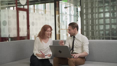 a red haired businesswoman has a relaxed meeting with a young man with a laptop on the couch in the common areas of the office building