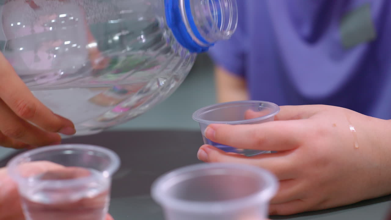 primer plano de agua que se vierte de una gran botella de plástico en pequeños vasos transparentes, con las manos sosteniendo los vasos y las gotas de agua visibles en la piel