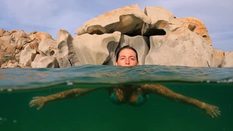 Half-underwater-scene-of-woman-swimming-toward-camera-at-Cala-Della-Chiesa-lagoon-with-eroded-granitic-rocks-in-background-of-Lavezzi-island-in-Corsica,-France