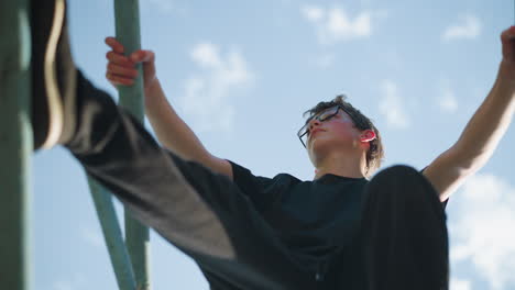 an upward view of a young boy sitting on outdoor blue iron equipment, holding onto the structure with both hands as he looks to the right