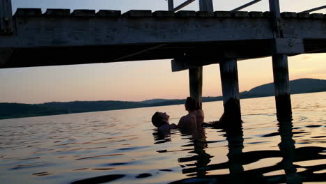 couple swimming under a pier at sunset