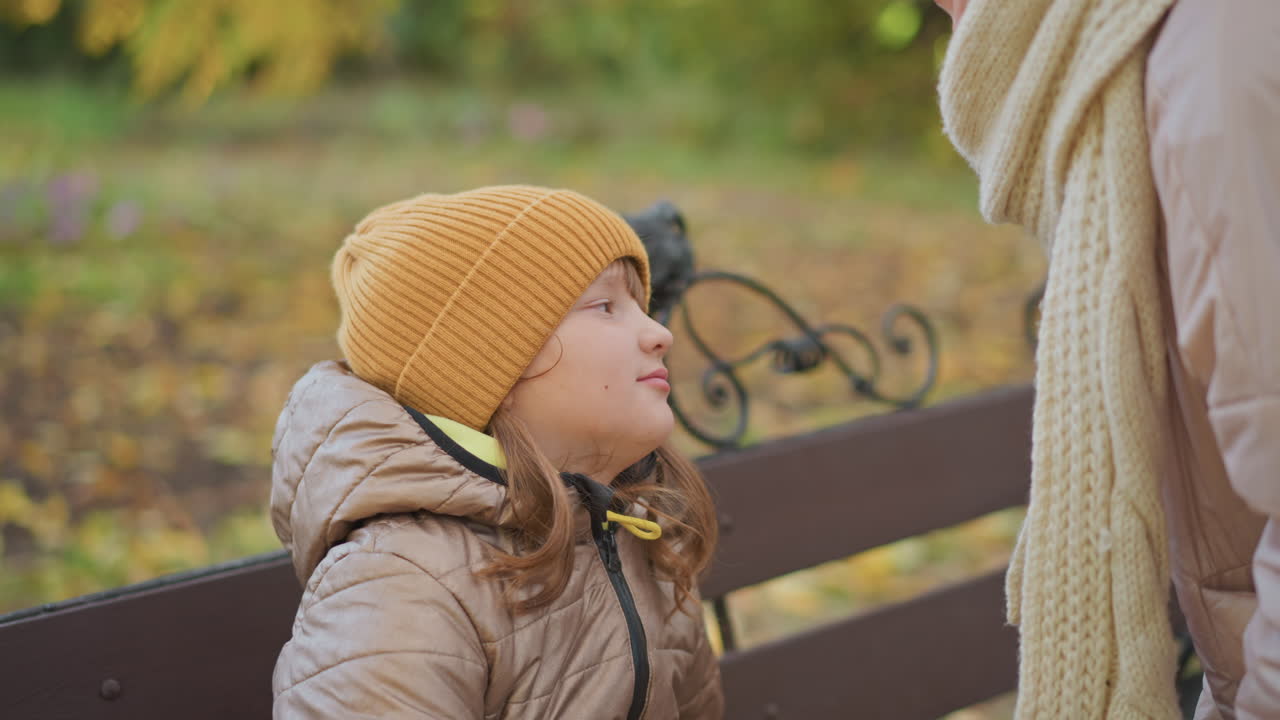 mother gently plays with daughter hair while leaning forward to kiss her on nose, captured in warm autumn setting with soft jackets and fallen leaves in peaceful park atmosphere